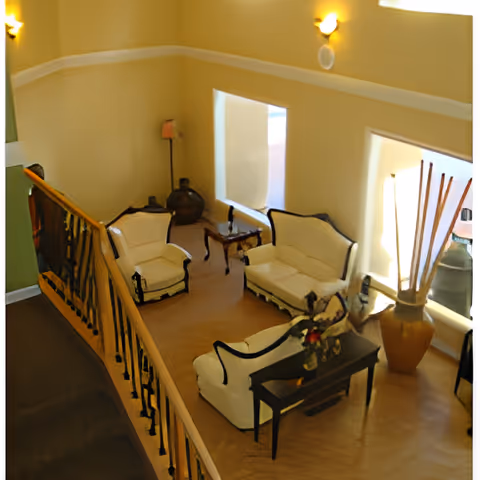 A cozy living room area with cream-colored walls and wooden flooring, featuring a white upholstered loveseat, two matching armchairs, a small wooden coffee table, and a side table with decorative items. The room is illuminated by wall-mounted lights and natural light from two windows. A large vase with tall decorative sticks is placed near one window, and a wooden railing borders the space.