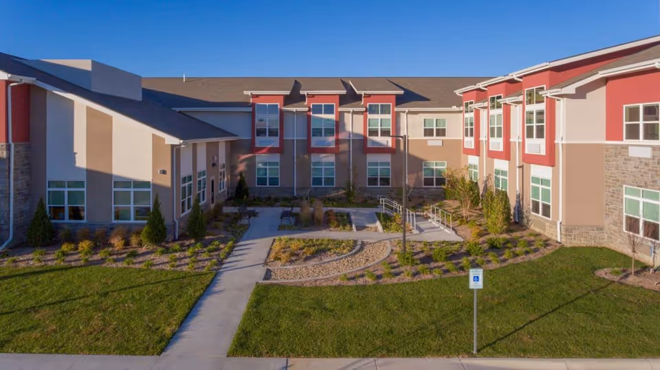 Exterior view of a modern two-story senior living building with a landscaped courtyard, walkways, and an accessibility ramp.