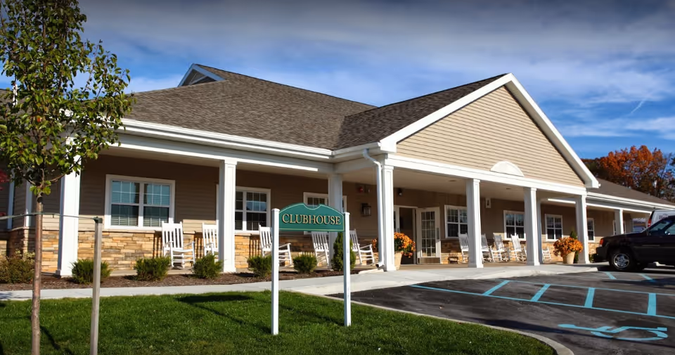 Front exterior of a clubhouse with rocking chairs and a parking lot.