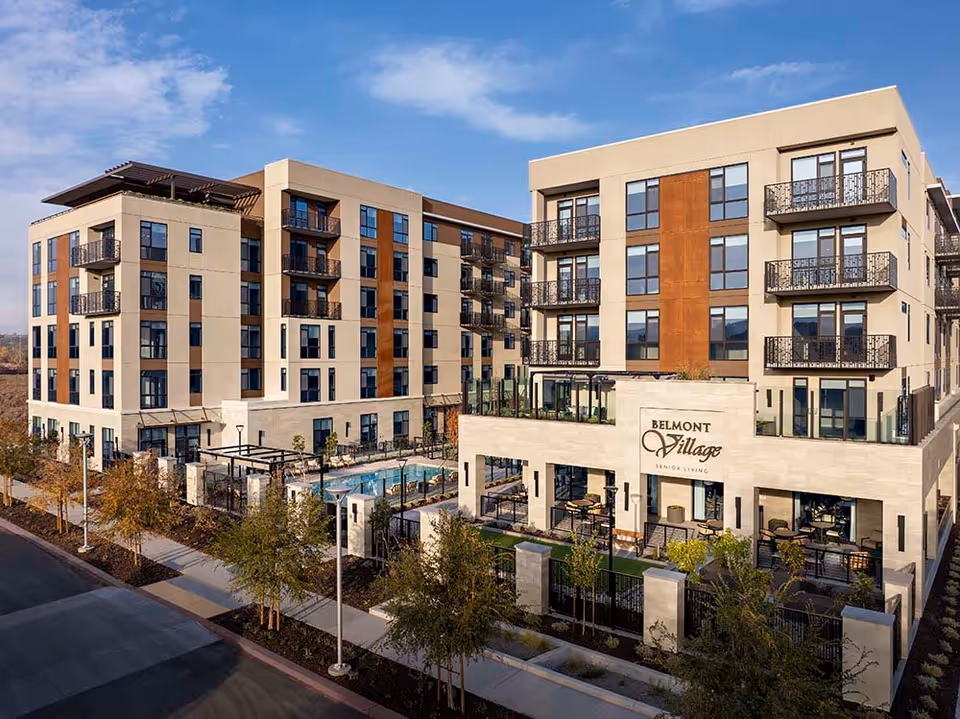 Modern multi-story senior living building with balconies, outdoor pool, and a visible 'Belmont Village' sign.