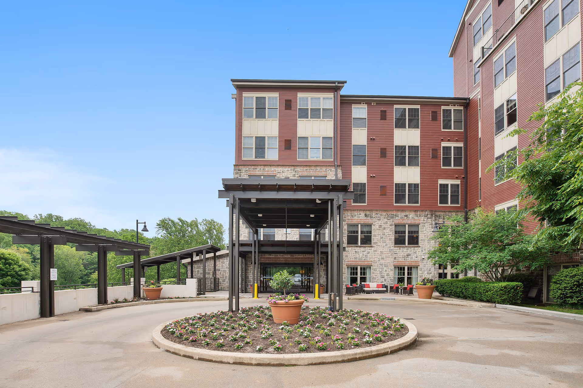 Front exterior view of Plush Mills Senior Living facility showing a multi-story building with red and beige siding, stone accents, a covered entrance, and a circular flower bed in the driveway under a clear blue sky.