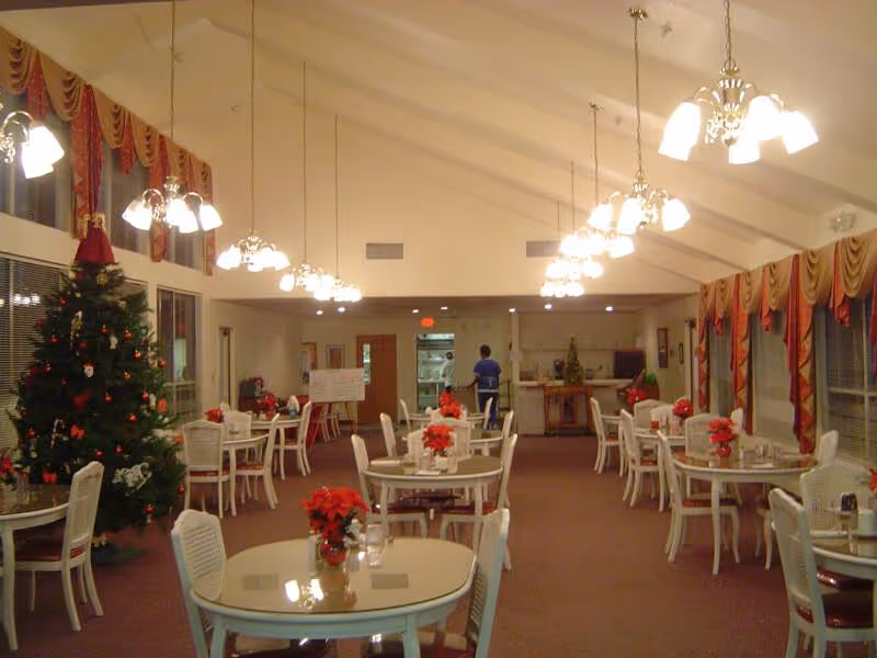 Spacious assisted-living dining room with round tables and chairs, chandeliers, holiday decorations including a Christmas tree, and a staff member near the serving area.