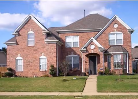Two-story red-brick suburban house with a front lawn, walkway, and blue sky.