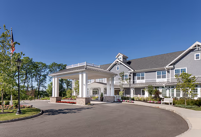 Exterior view of a senior living facility building with a covered entrance, landscaped garden, and clear blue sky.
