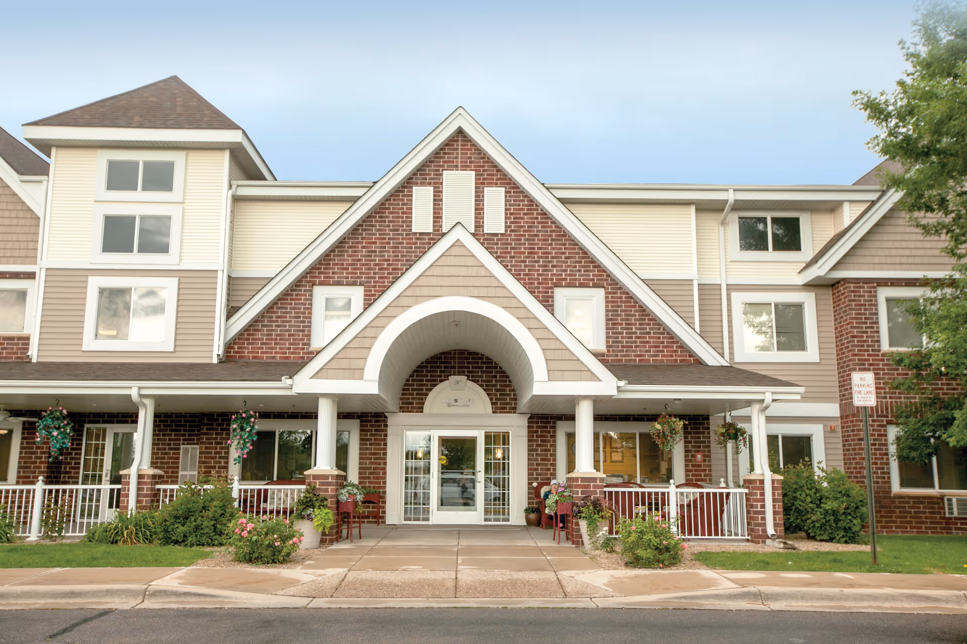 Front exterior view of a senior living facility building with a peaked roof entrance, brick and beige siding, white columns, hanging flower baskets, and a paved walkway leading to glass double doors.
