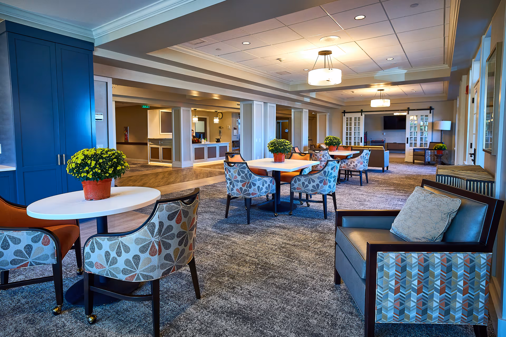 Spacious senior living lounge with round tables, patterned chairs, and potted plants.