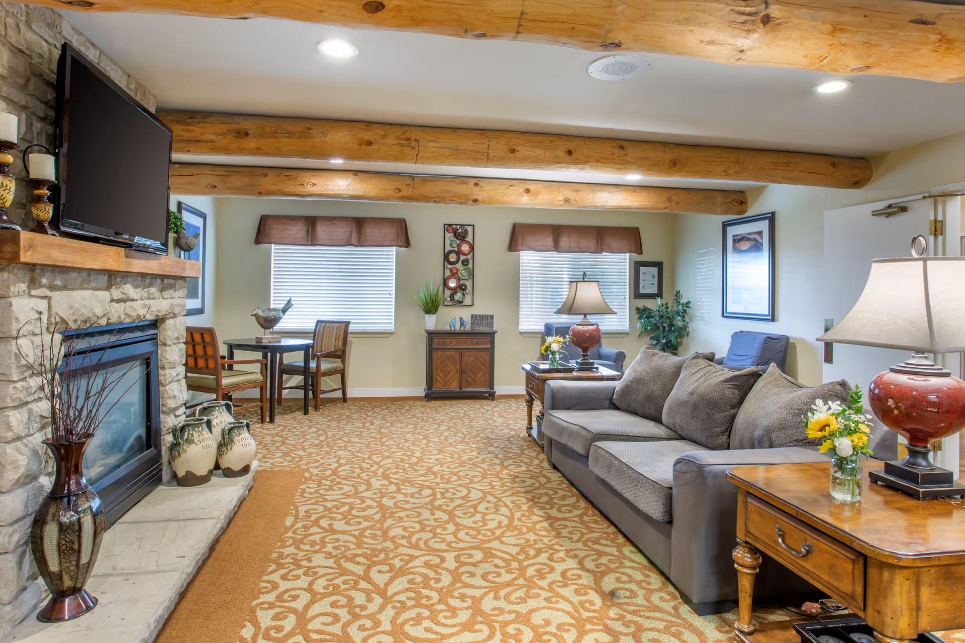Comfortable living room with a sofa, fireplace and TV, small dining table, decorative wooden beams, and lamps.