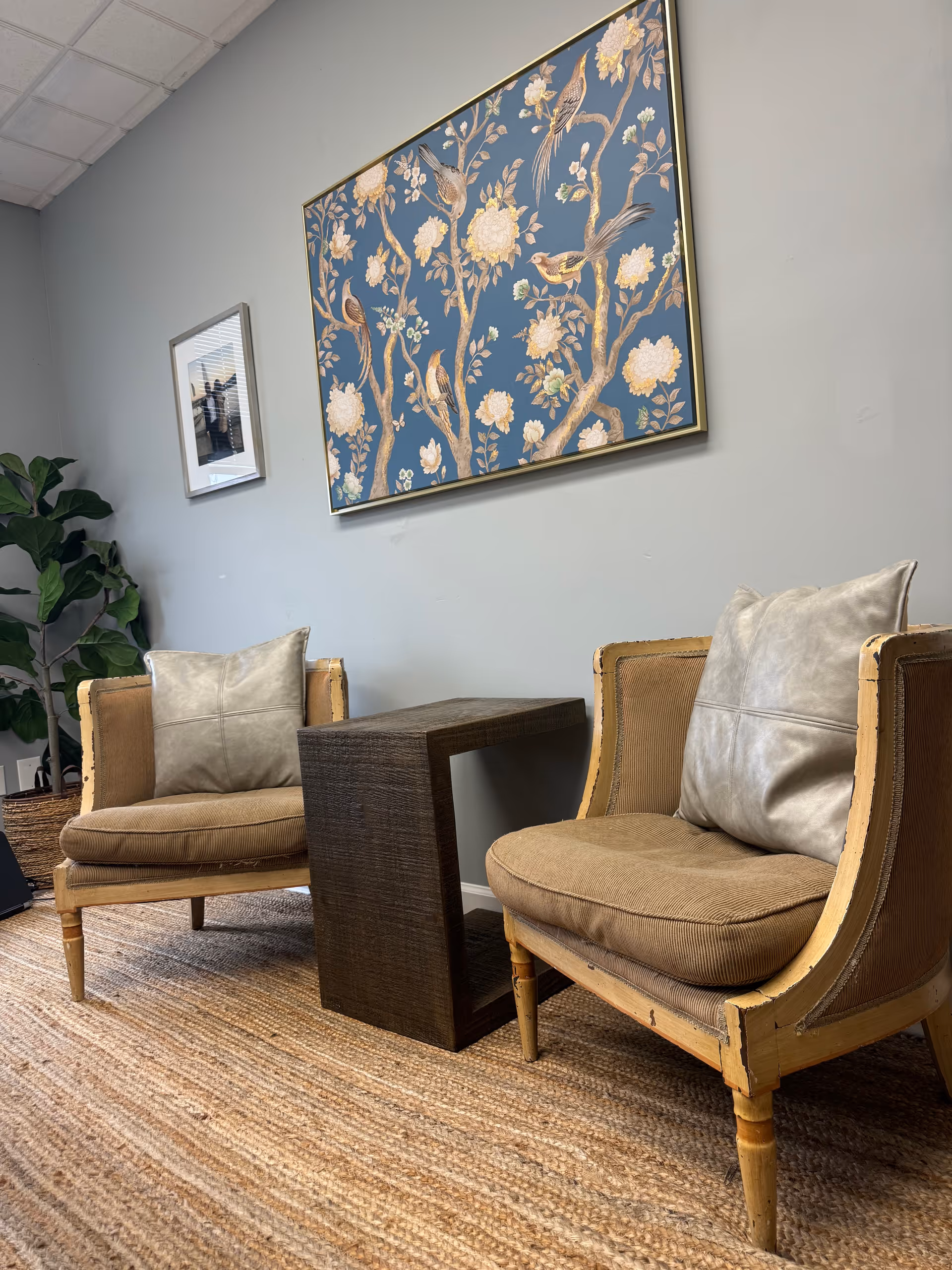 Two beige chairs flank a dark side table in a lounge with floral wall art.