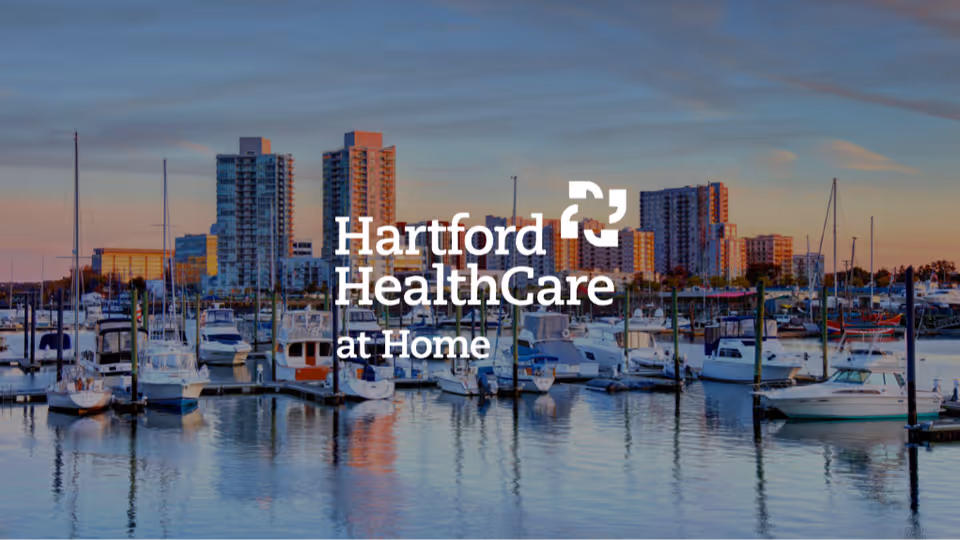 Marina with boats and a city skyline, overlaid by Hartford HealthCare at Home logo.