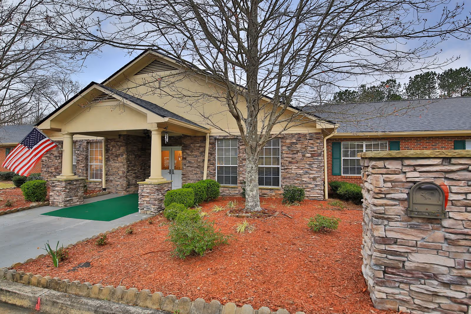 Front exterior view of Manchester Court Assisted Living and Memory Care building with stone and brick facade, a covered entrance with columns, an American flag, and landscaped garden with mulch and shrubs.