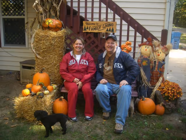 A man and a woman sitting on a bench outside a house decorated for fall with pumpkins, hay bales, and two scarecrows. A small black dog stands on the grass in front of them. A wooden sign above the bench reads 'Welcome to Family Living'.