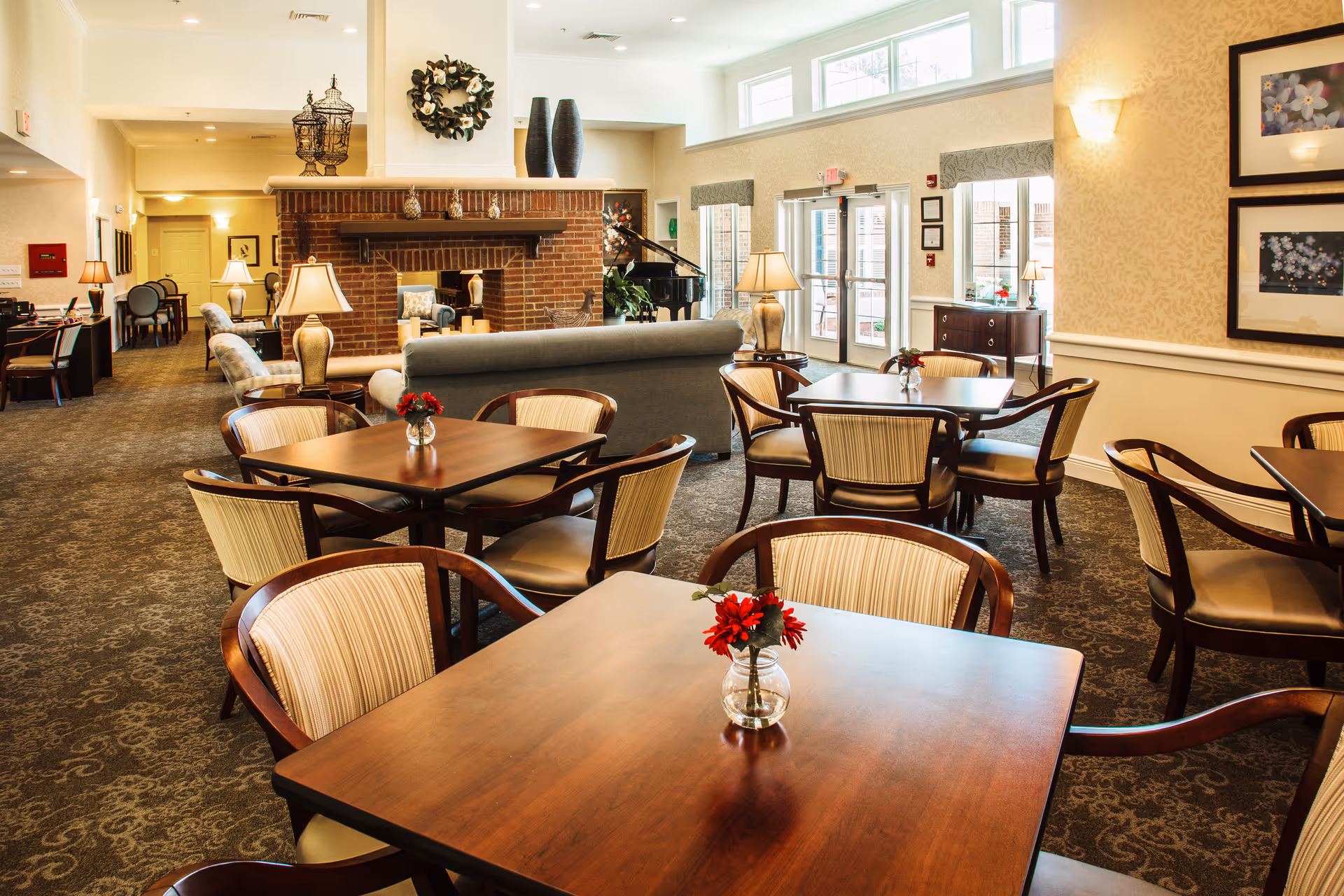 A bright and spacious senior living facility common area with several wooden tables and chairs arranged for dining or socializing. Each table has a small vase with red flowers. In the background, there is a brick fireplace with decorative items on the mantel, a gray sofa, and a grand piano near large windows and glass doors letting in natural light. The room has warm beige walls, patterned carpet, and framed artwork on the walls.