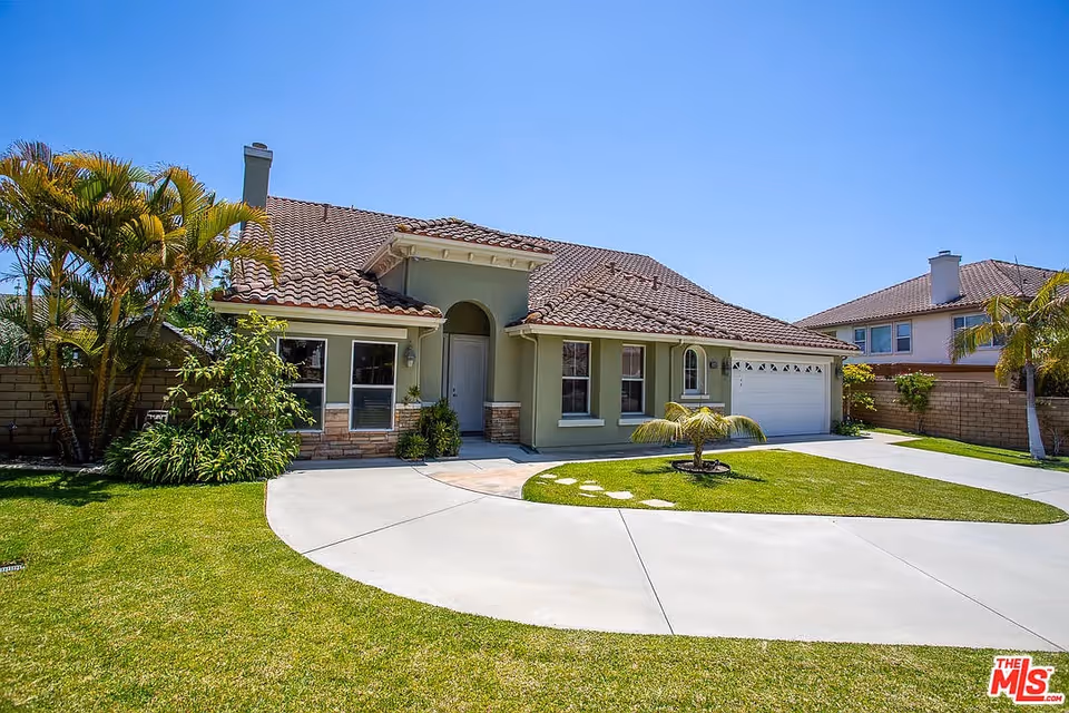 Single-story beige home with clay tile roof, curved driveway, green lawn, and palm trees.