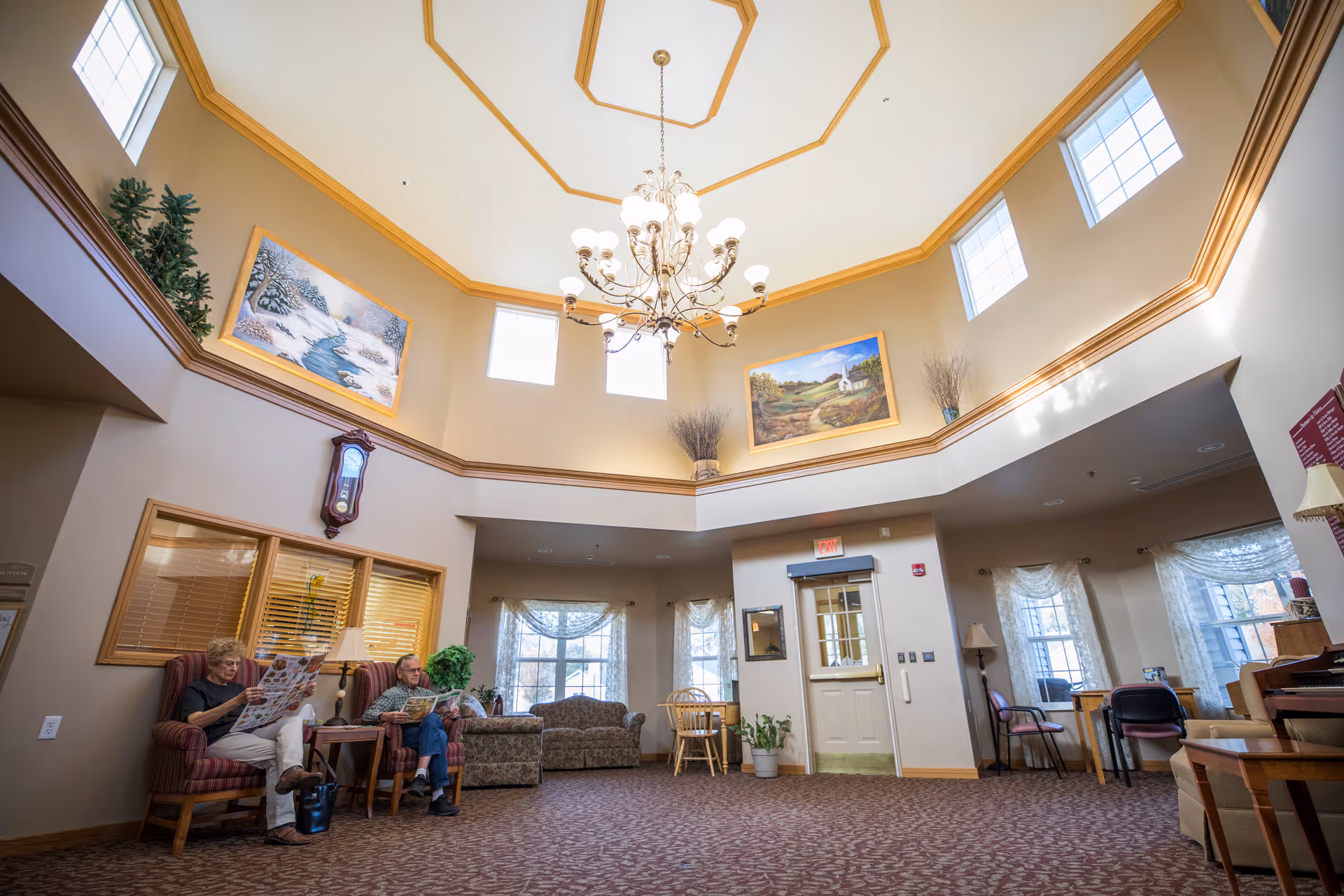 Sunlit senior living common room with a high domed ceiling and chandelier, seating areas, paintings, and two residents reading.