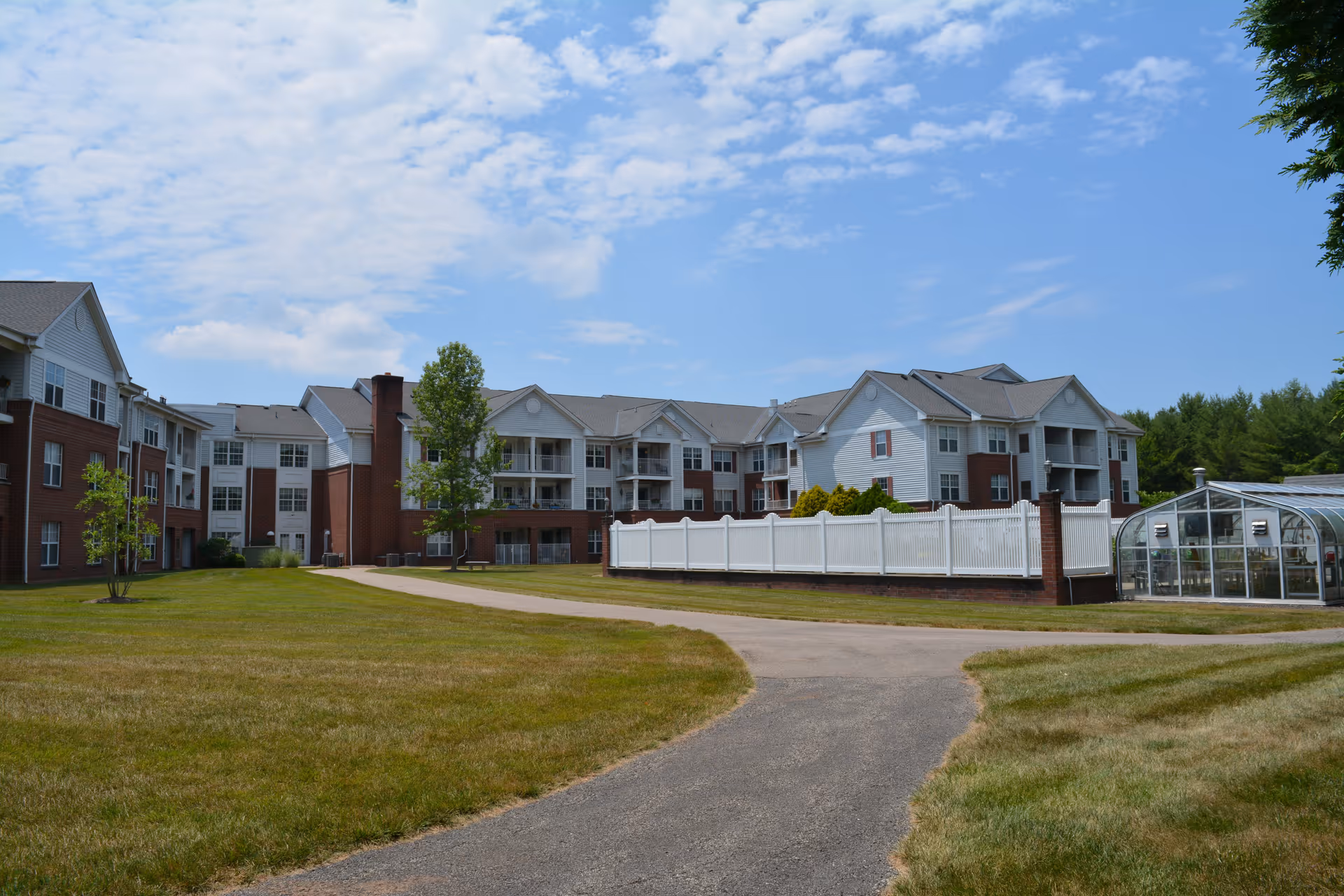 Exterior view of a senior living facility with a three-story building featuring white and red brick walls, balconies, and a white fence surrounding a small greenhouse. The scene includes a paved pathway and a grassy lawn under a partly cloudy blue sky.