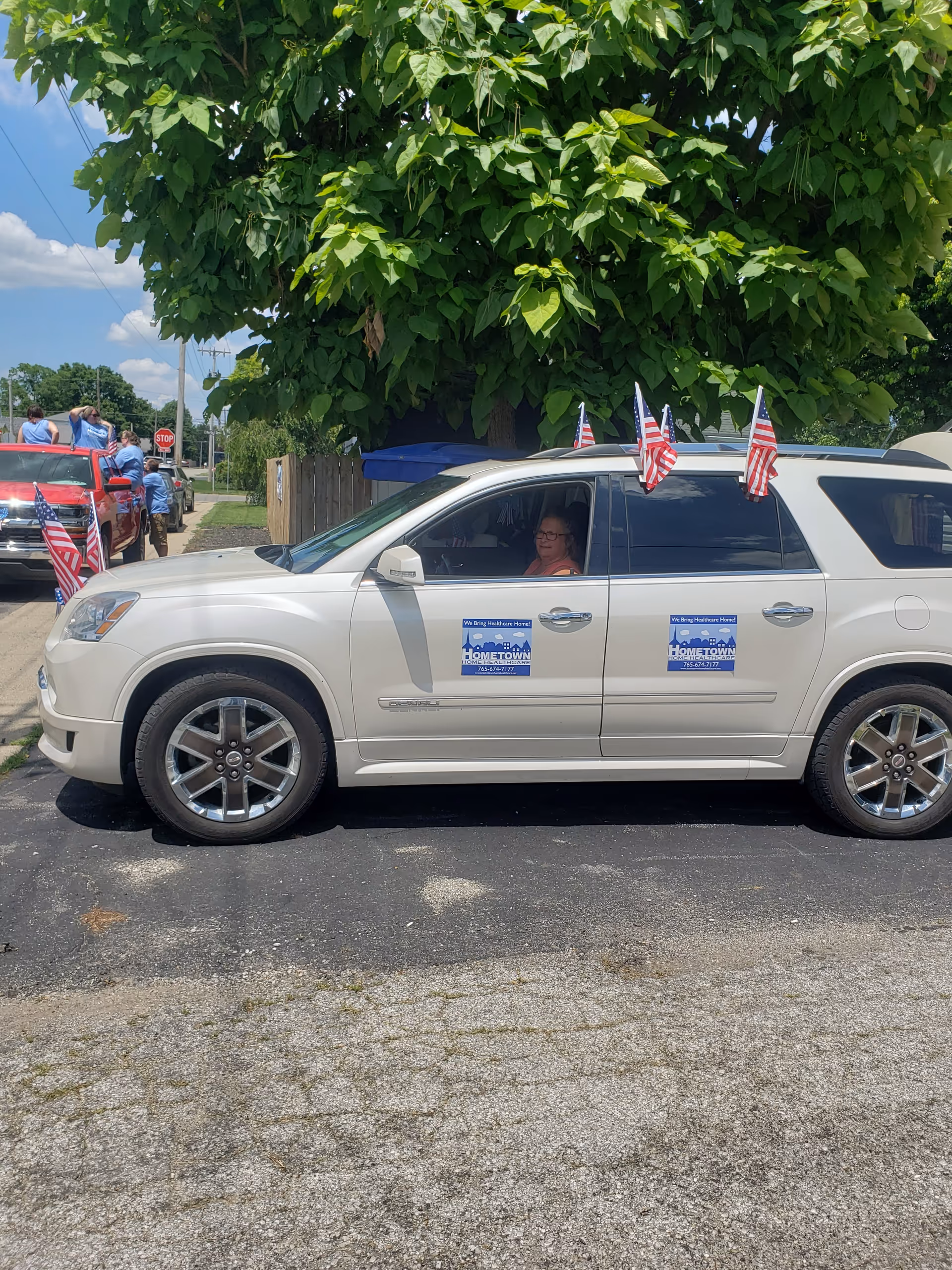 White SUV with Hometown Home Healthcare stickers and small flags parked outdoors beneath a large tree.