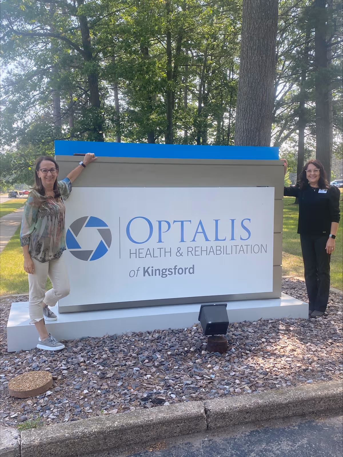 Two women pose beside a large Optalis Health & Rehabilitation of Kingsford sign outdoors.
