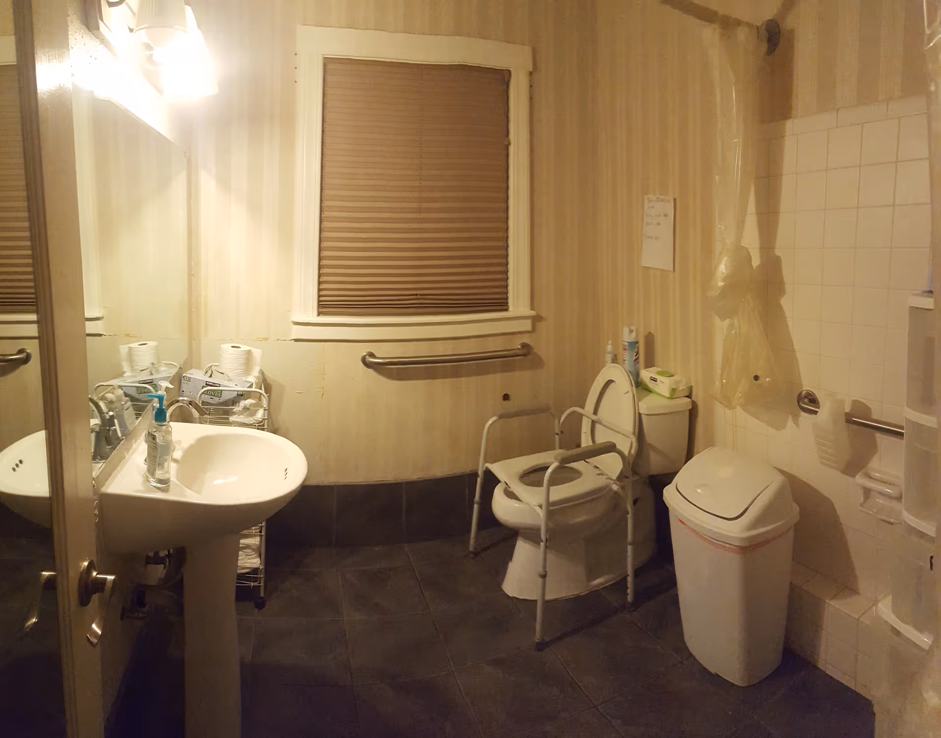 A bathroom with a pedestal sink, a mirror above it, a window with closed blinds, a toilet with safety rails around it, a trash can, and a shower area with a curtain. The walls have vertical striped wallpaper and there are grab bars installed near the toilet and shower for accessibility.