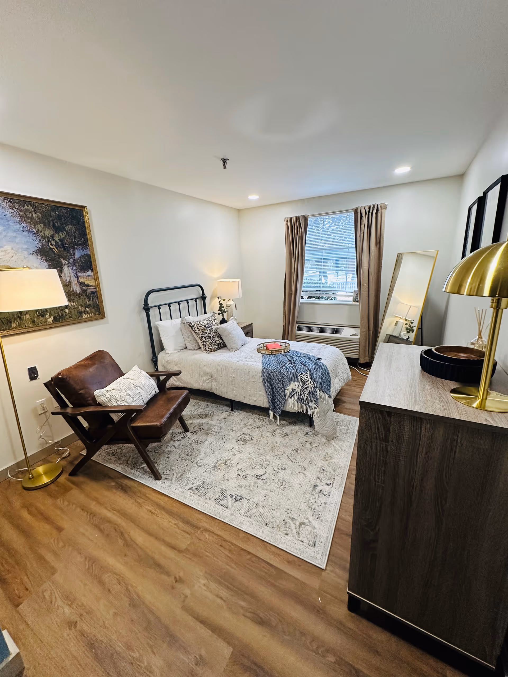 A cozy senior living bedroom with a single bed dressed in white bedding and decorative pillows, a blue throw blanket, a wooden armchair with a white pillow, a floor lamp, a framed painting on the wall, a window with beige curtains, a full-length mirror, and a wooden dresser with a gold lamp and decorative items.