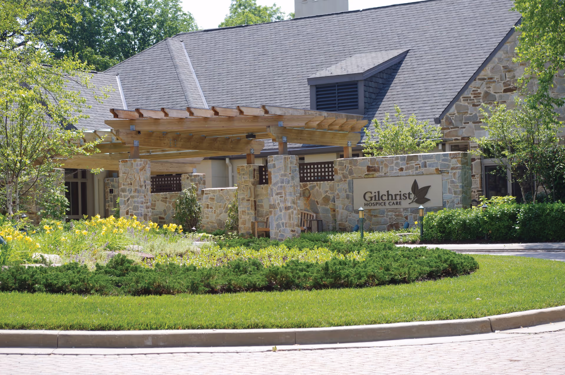 Stone exterior building with shaded entrance and Gilchrist Hospice Care sign amid landscaping.