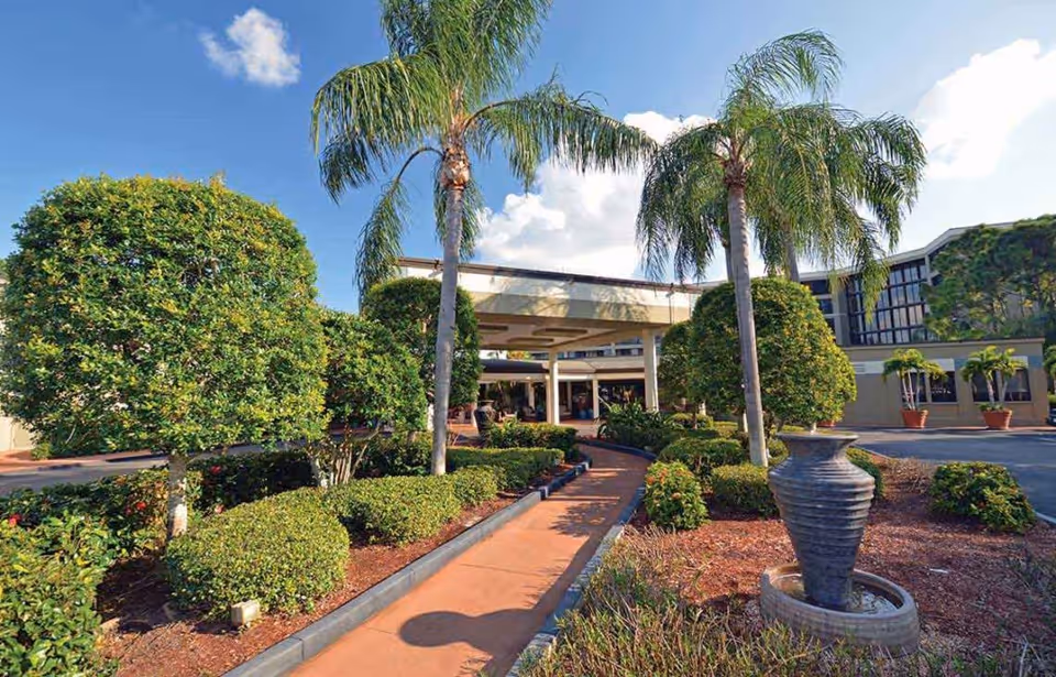 Covered entrance of a senior living building with palm trees, manicured hedges, a walkway, and a decorative urn fountain.