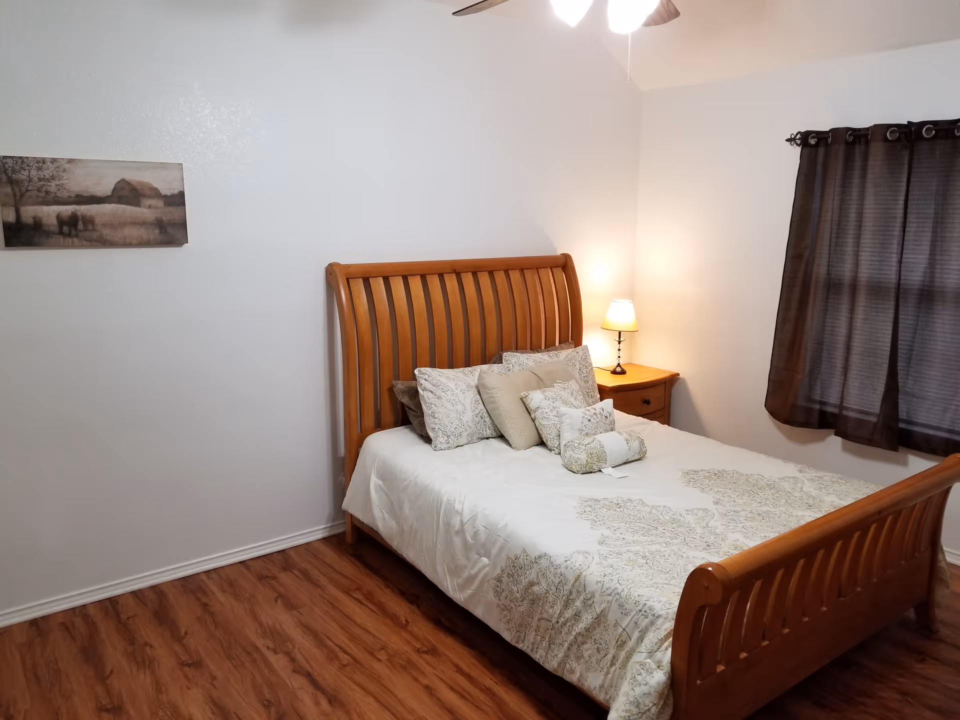Wooden bed with white bedding, pillows, nightstand, lamp, and dark curtains.