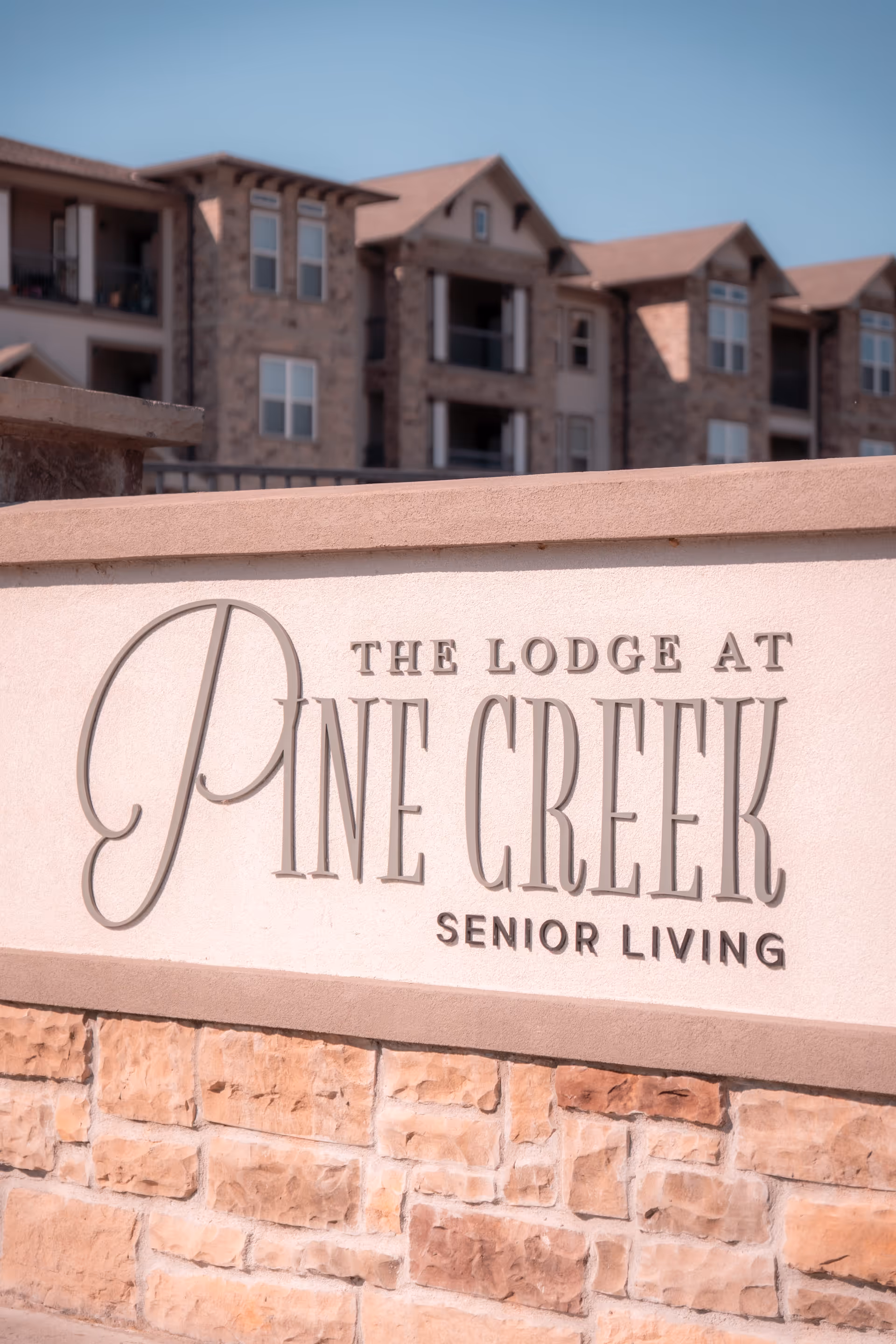 Close-up view of a stone and stucco sign that reads 'The Lodge at Pine Creek Senior Living' with a multi-story residential building in the background under a clear blue sky.