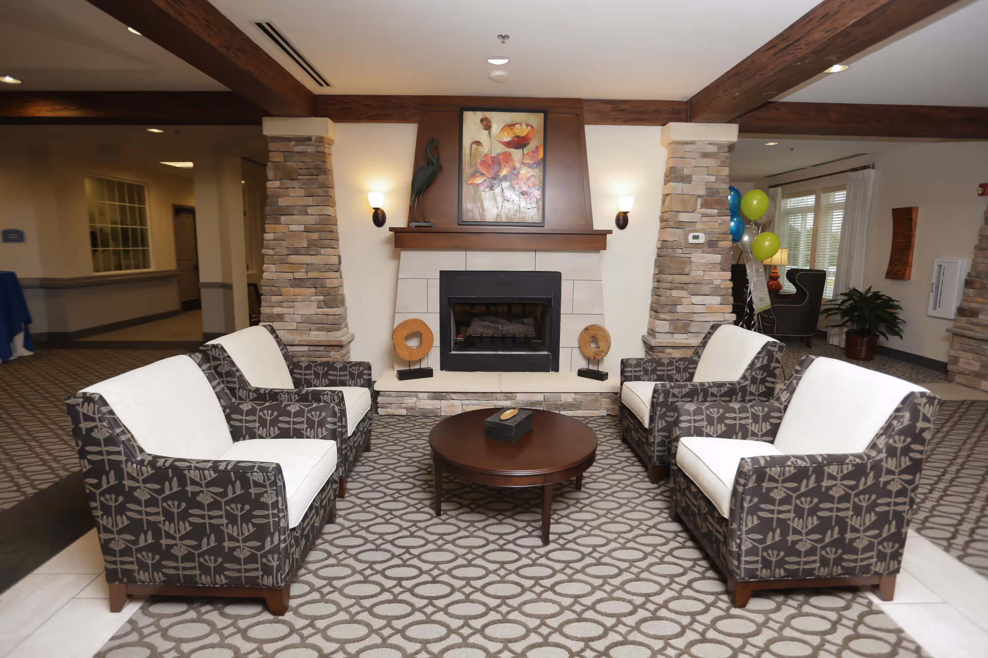 A communal living area with four patterned armchairs around a round coffee table facing a fireplace and artwork.