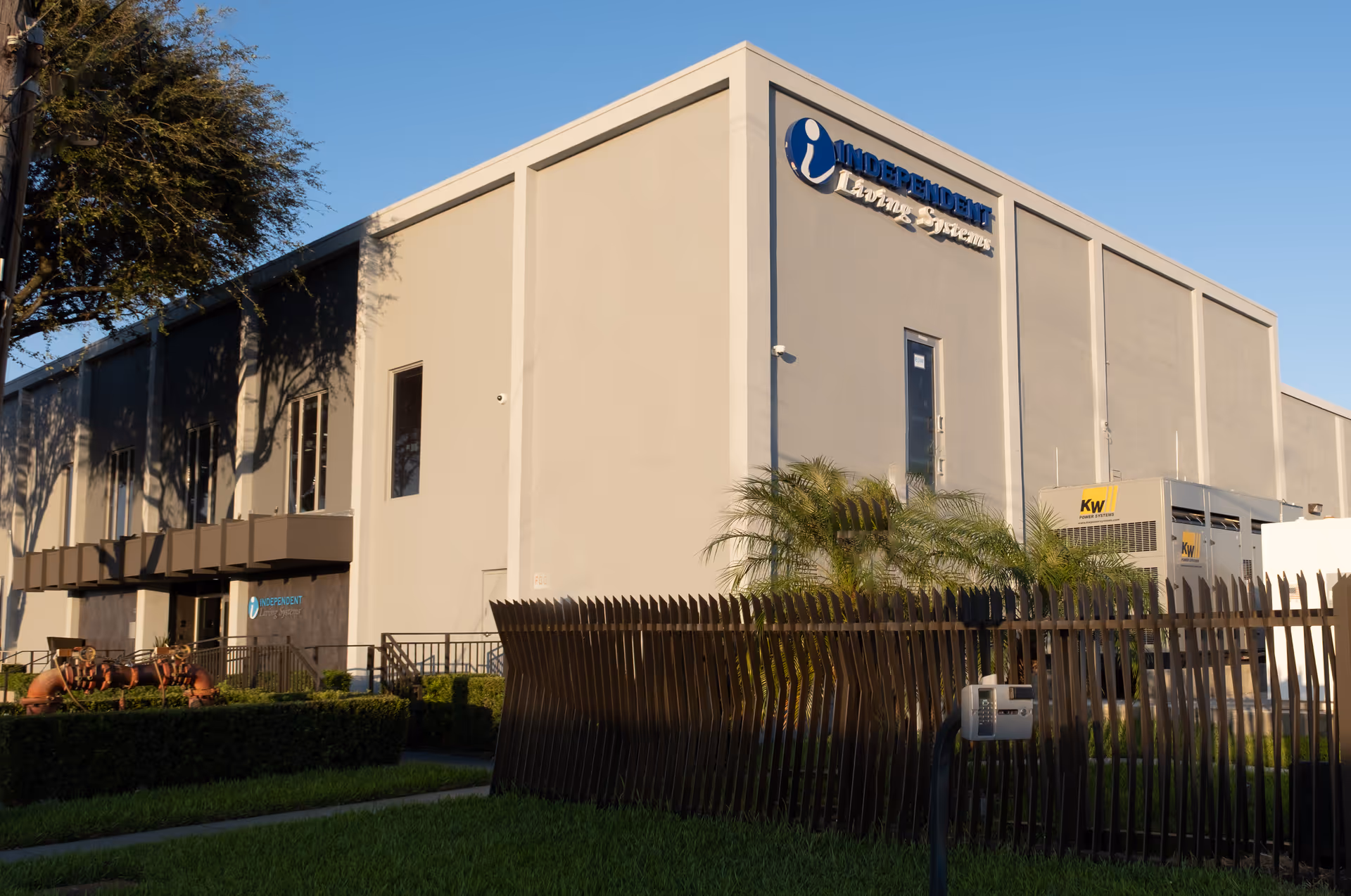 Front exterior of a beige Independent Living Systems facility with fencing and palm trees