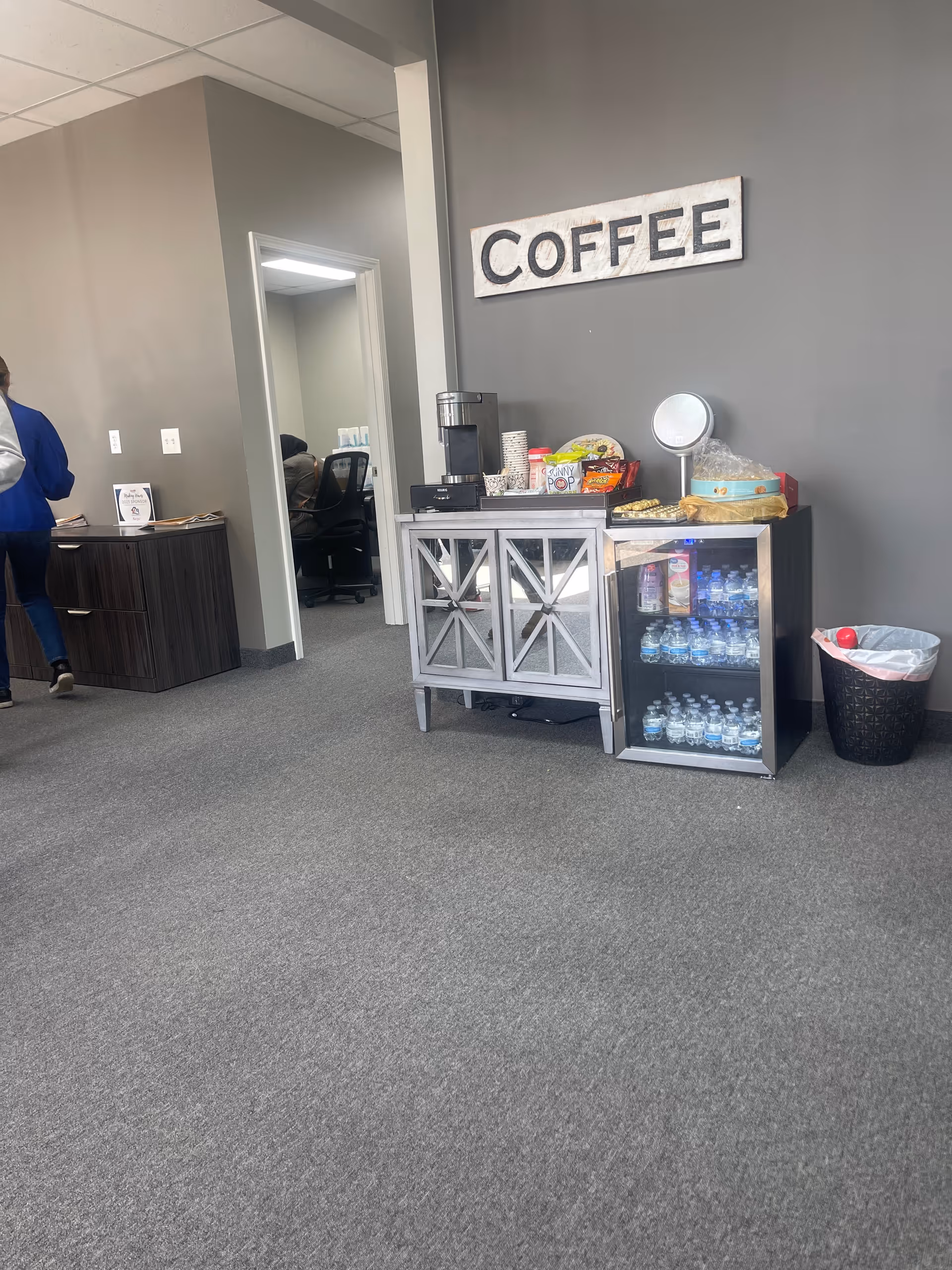 A coffee station with snacks and water bottles against a gray wall in a facility lobby.