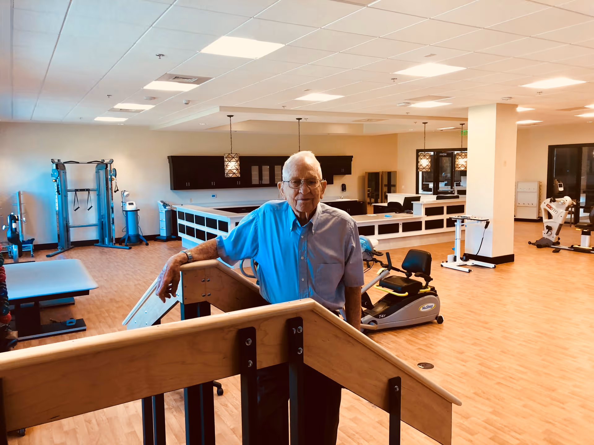 An elderly man wearing glasses and a light blue shirt stands in a spacious rehabilitation or physical therapy room with wooden flooring. The room contains various exercise equipment including a recumbent bike, weight machines, and parallel bars. The background shows a counter area with pendant lights and cabinets.