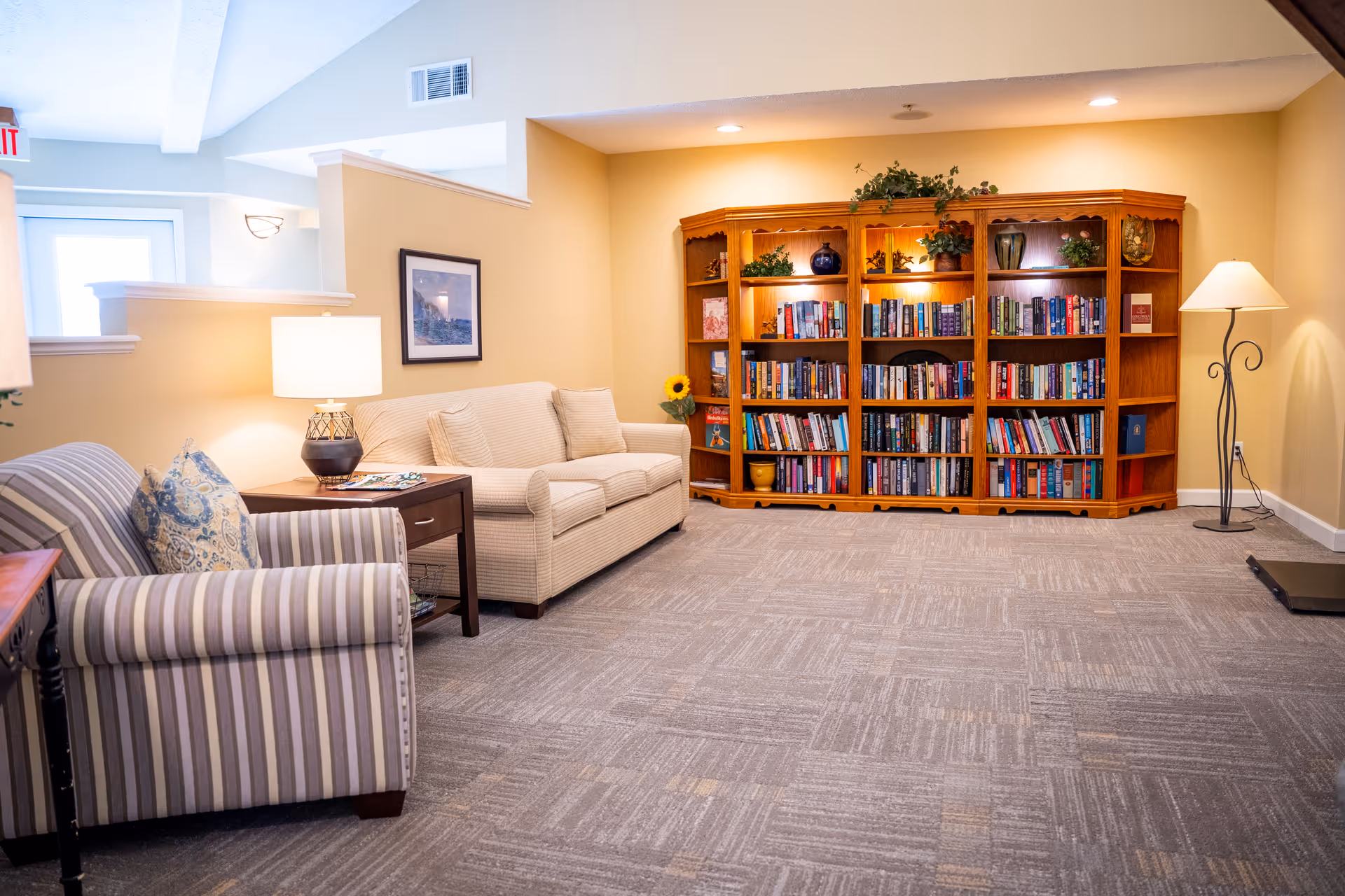 A cozy living room area with a beige couch and a striped armchair with a decorative pillow. There is a wooden side table with a lamp and magazines between the couch and armchair. In the background, a large wooden bookshelf filled with books and decorative items is against a yellow wall. A floor lamp stands to the right of the bookshelf, and the room has carpeted flooring.