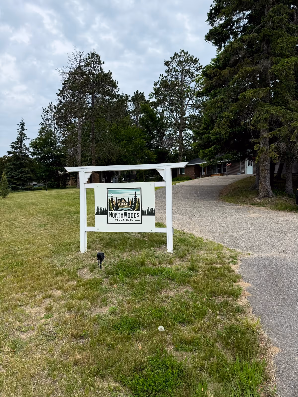 A white wooden signpost on a grassy area with the sign reading 'Northwoods Villa Inc.' The sign features an illustration of a house surrounded by pine trees. Behind the sign is a paved driveway leading to a building partially obscured by tall trees under a cloudy sky.