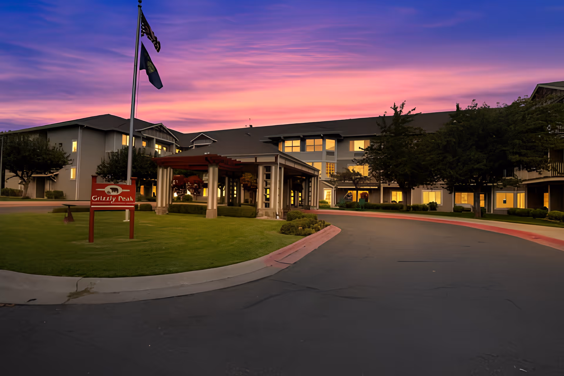 Exterior view of the Holiday Grizzly Peak senior living facility at sunset, showing a large building with multiple windows lit from inside, a covered entrance, a curved driveway, green lawn, trees, and two flagpoles with flags.