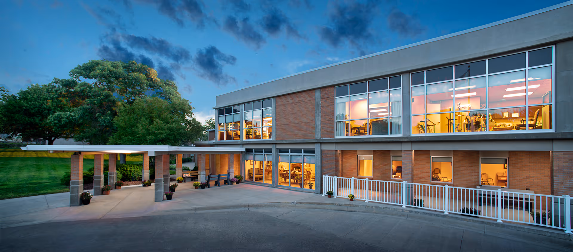 Exterior view of a two-story brick building at dusk with large windows showing interior lighting. The building has a covered entrance with columns and a paved driveway. There are green trees and grass surrounding the building under a partly cloudy sky.
