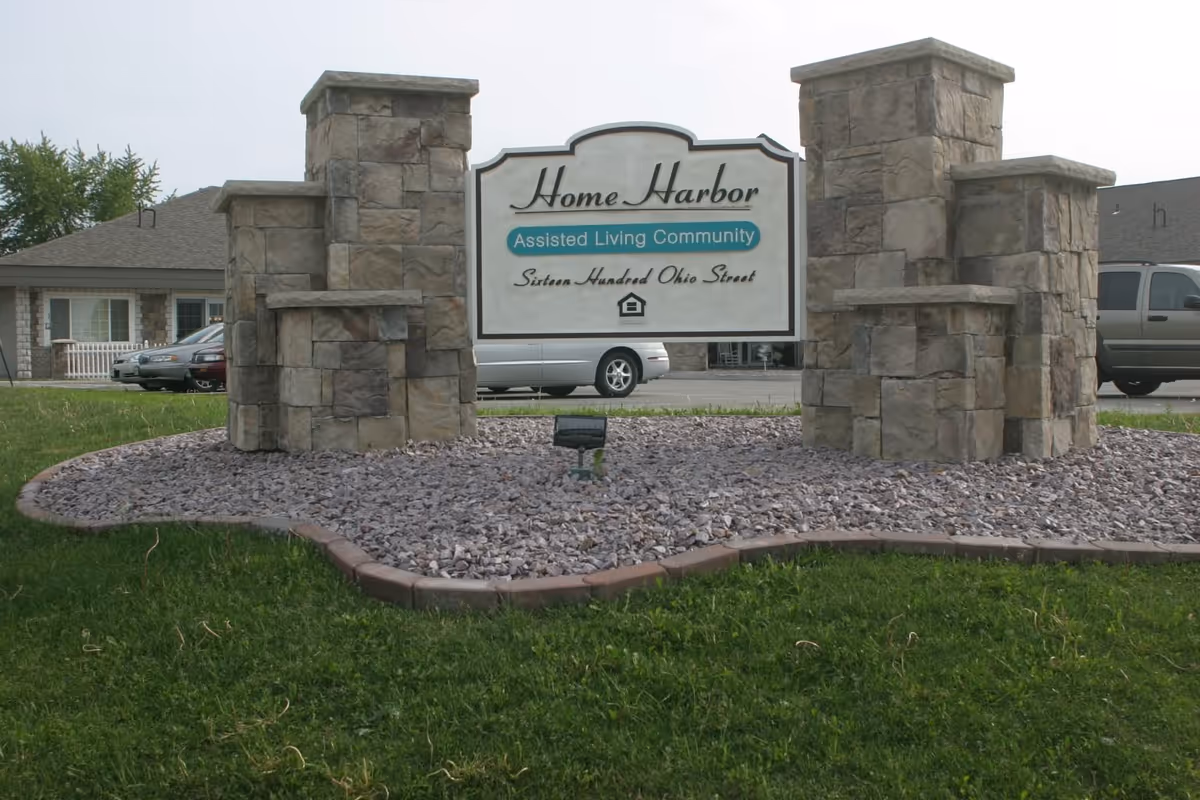 Stone monument sign for Home Harbor Assisted Living Community with the address Sixteen Hundred Ohio Street, set in a landscaped area with grass and rocks, with buildings and parked vehicles in the background.