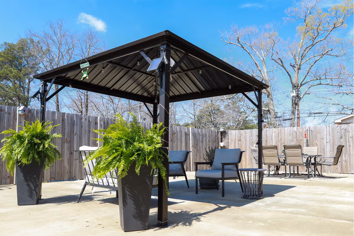 Outdoor patio with a metal gazebo, seating, dining table, and large potted ferns next to a wooden privacy fence under a blue sky.