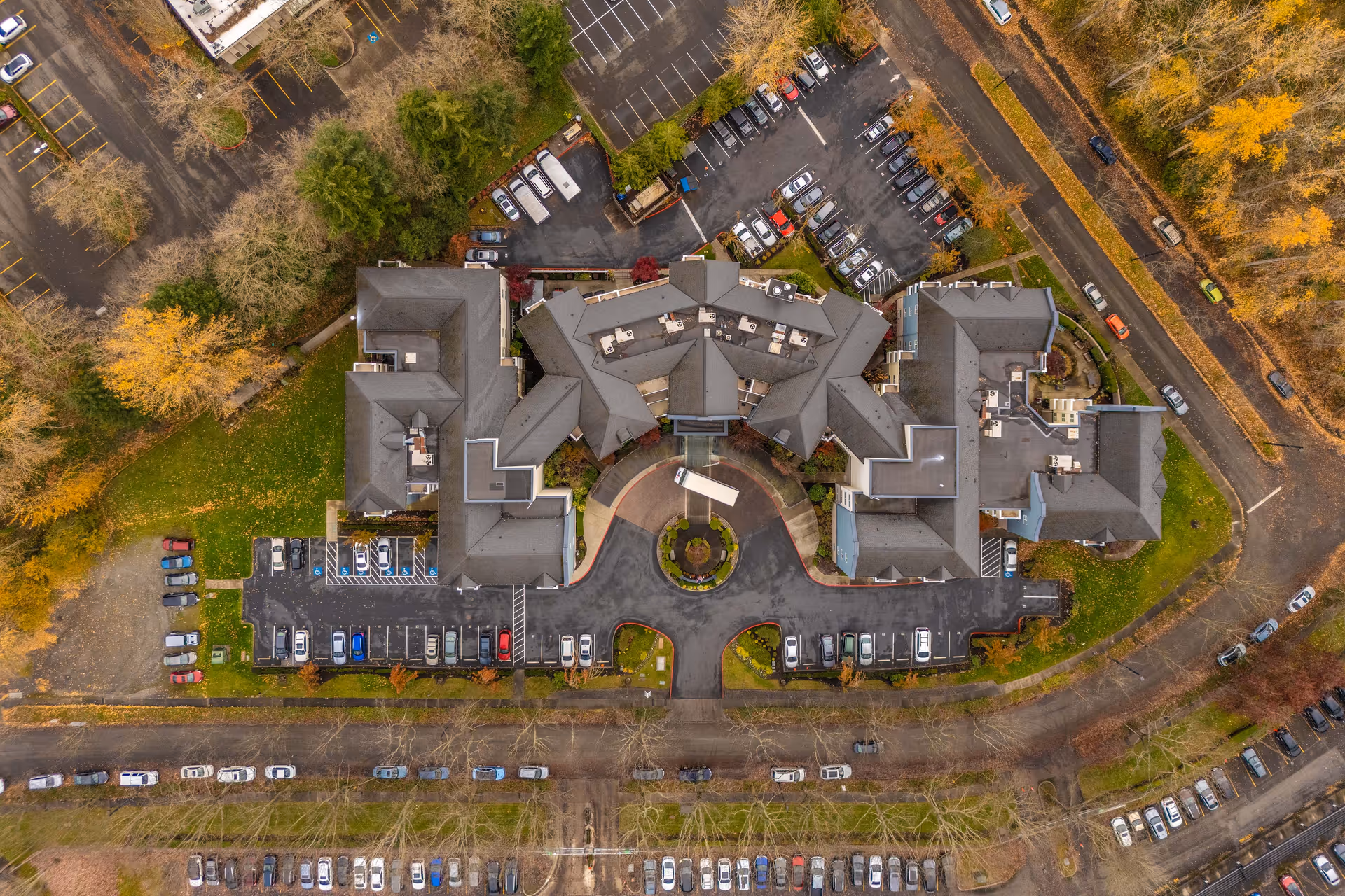 Aerial view of Chateau Valley Center Retirement facility showing a large building complex with multiple wings, surrounded by parking lots filled with cars, trees with autumn foliage, and adjacent roads.