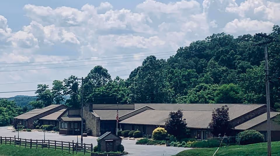Exterior view of a single-story building with a sloped roof surrounded by greenery and trees under a partly cloudy sky. There is a paved driveway and a wooden fence in the foreground.