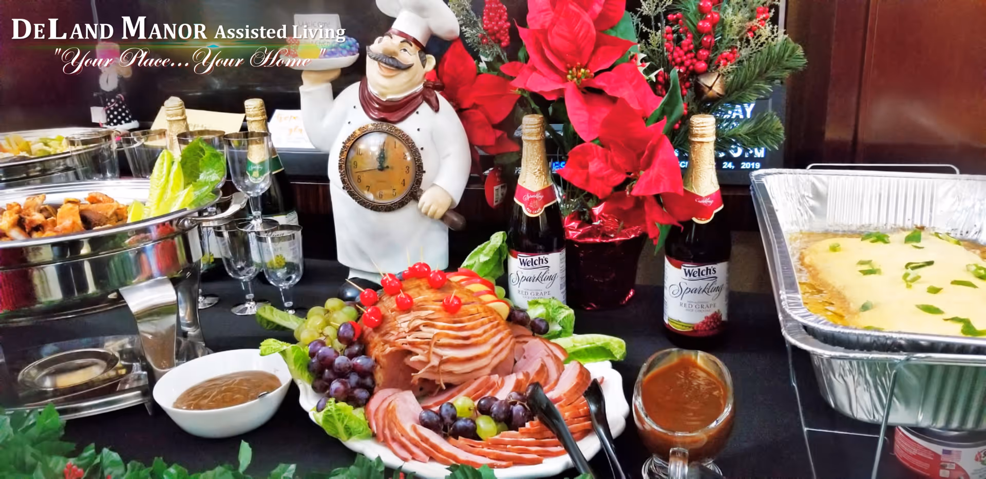 A buffet table at DeLand Manor Assisted Living featuring a sliced ham garnished with grapes and cherries, a bowl of gravy, a tray of cooked food, bottles of Welch's Sparkling juice, a decorative chef figurine with a clock on its belly, and a festive poinsettia plant.