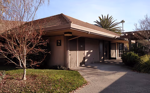 Single-story senior care facility exterior with shaded entrance, landscaping, and a tall palm tree.