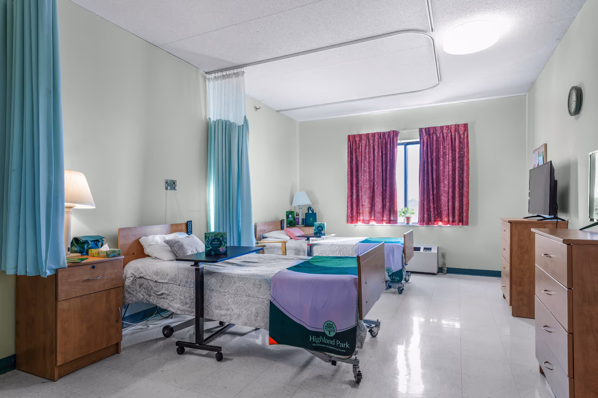 A bright multi-bed patient room with privacy curtains, furniture, and a mounted TV.
