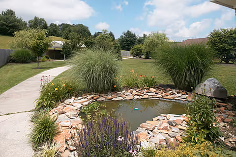 Outdoor garden with a stone bordered pond, walkways, and ornamental grasses.