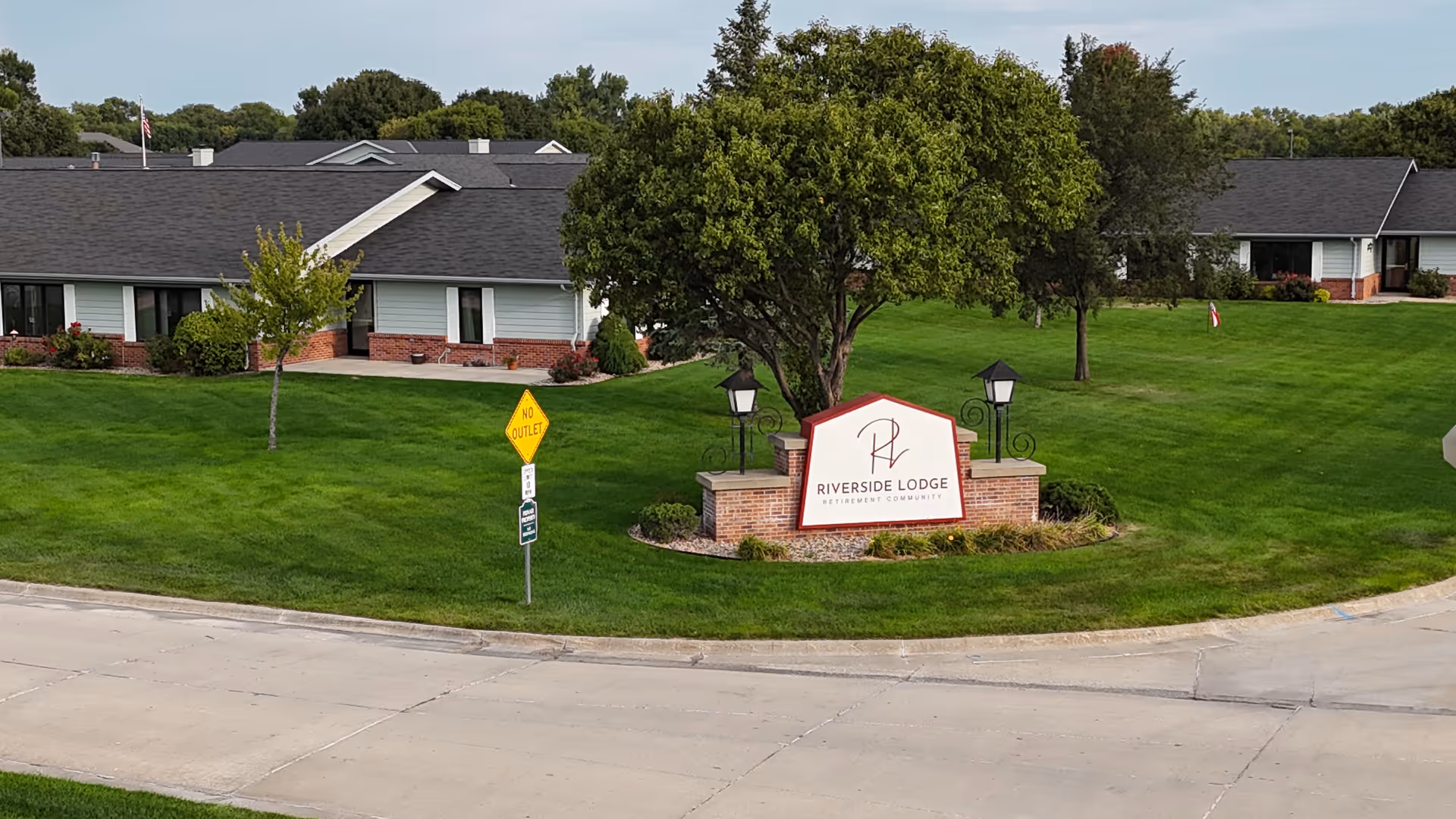 Exterior view of Riverside Lodge Retirement Community showing a well-maintained green lawn with a large tree and a brick sign displaying the facility's name. Single-story buildings with dark roofs and white siding are visible in the background under a partly cloudy sky.