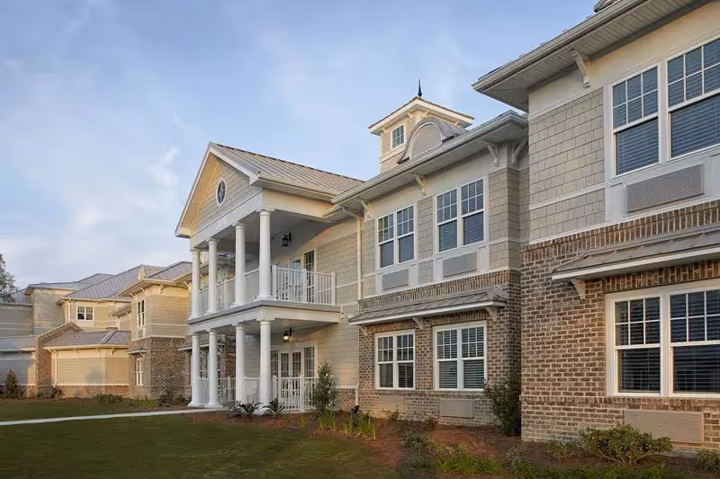 Exterior front of a two-story senior living building with columns, balconies, brick and siding facade, and a small landscaped lawn.