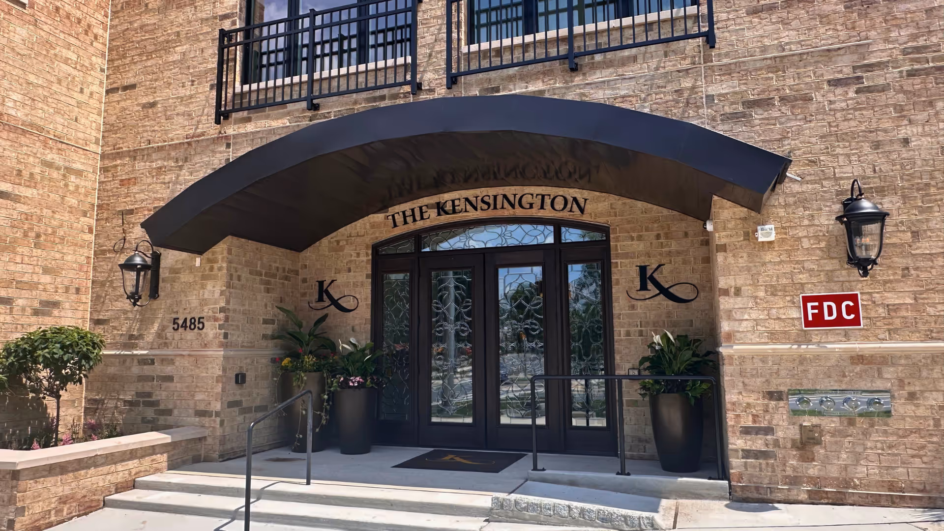 Brick building entrance with arched canopy, double doors, and potted plants at The Kensington Bethesda.