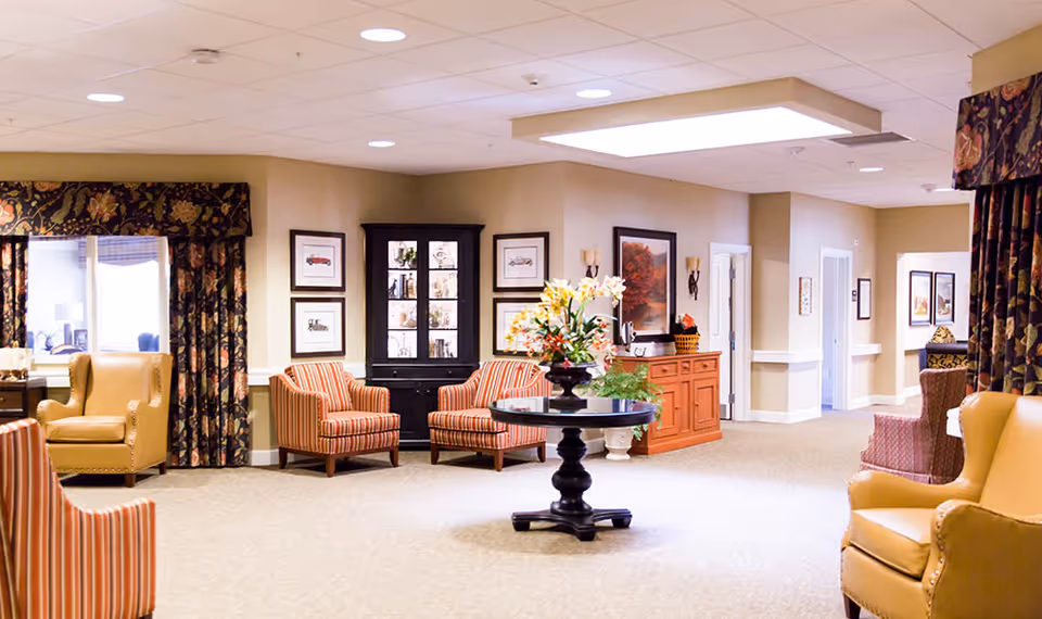 A well-lit common area in a senior living facility featuring comfortable armchairs and striped chairs arranged around a central round table with a floral arrangement. The walls are decorated with framed artwork, and there are large windows with patterned curtains allowing natural light to enter. The space has a warm and inviting atmosphere with beige walls and carpeted floors.