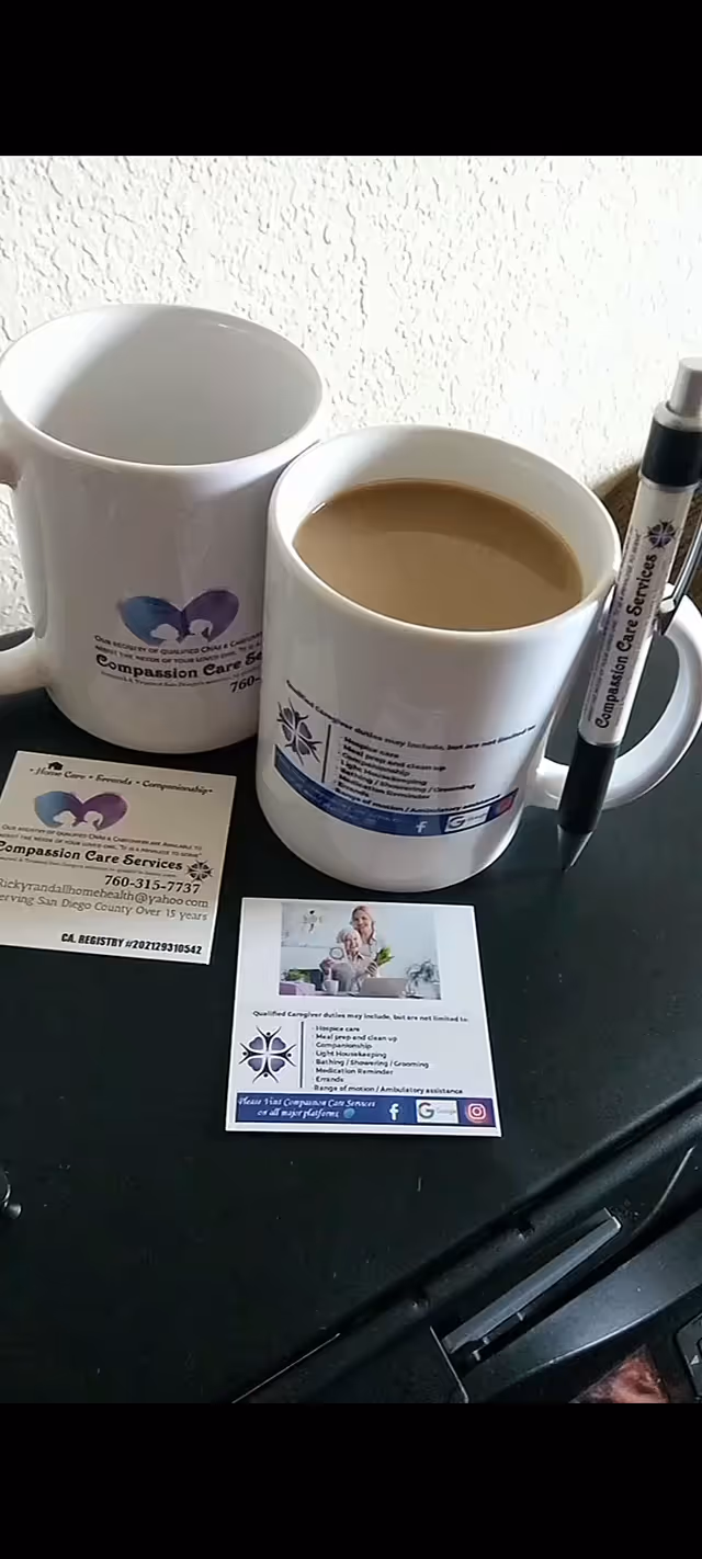 Two white mugs with Compassion Care Services logos beside a pen and business cards on desk.