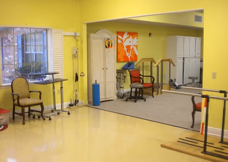 Interior room with yellow walls and a large window showing an outside view. The room contains chairs, a small table, a white cabinet, exercise equipment including parallel bars and a blue foam roller, and a colorful orange and white wall art. The floor is a combination of smooth yellow and carpeted areas.
