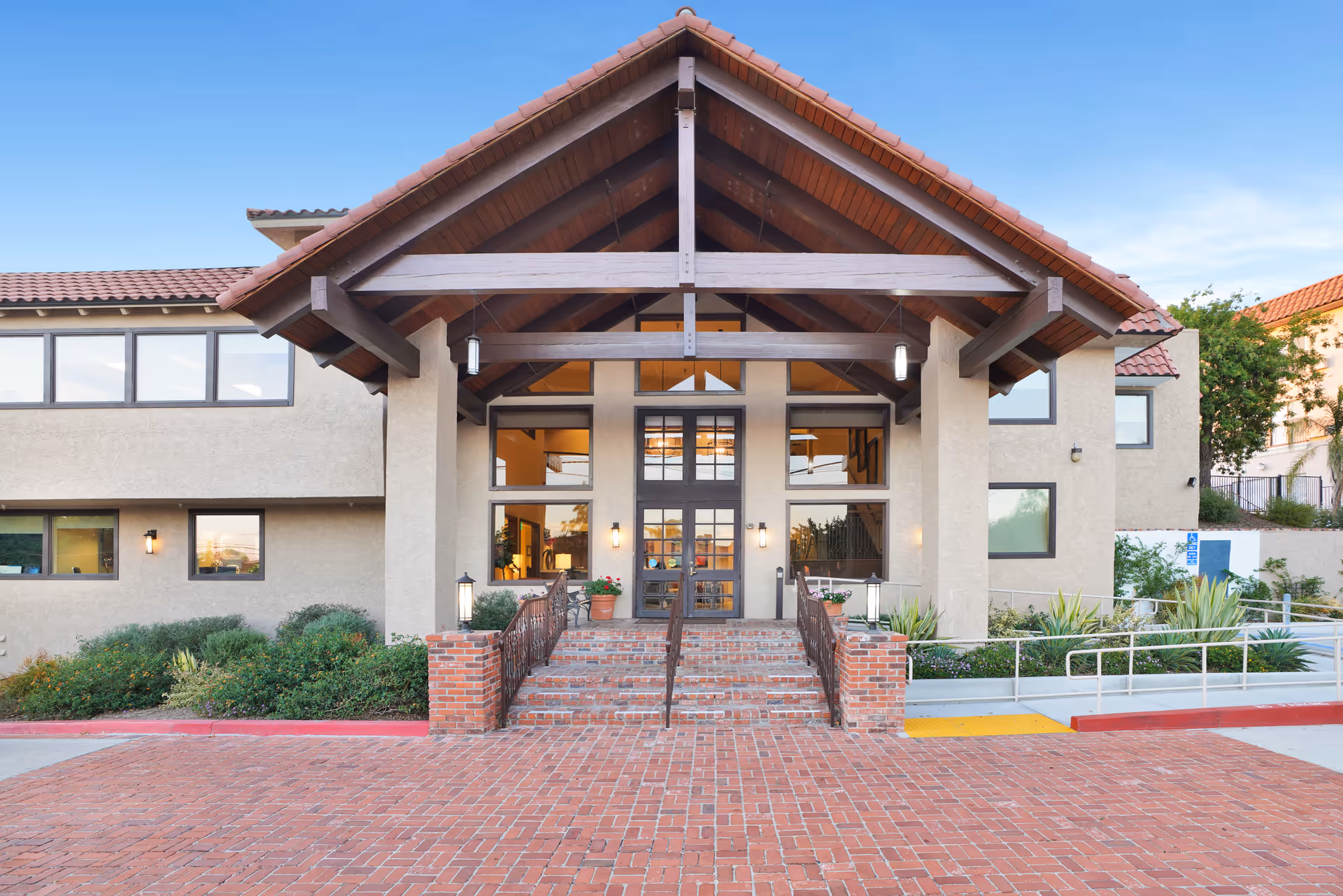 Front exterior view of The Montera facility showing a building entrance with a large wooden gabled roof supported by columns, brick steps leading up to double glass doors, and surrounding landscaping with bushes and plants.